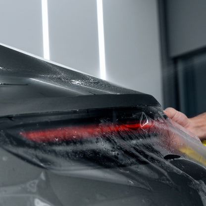 A close-up of a person applying a clear protective film or wrap to the rear quarter panel and taillight area of a dark gray car, with water droplets visible on the surface.