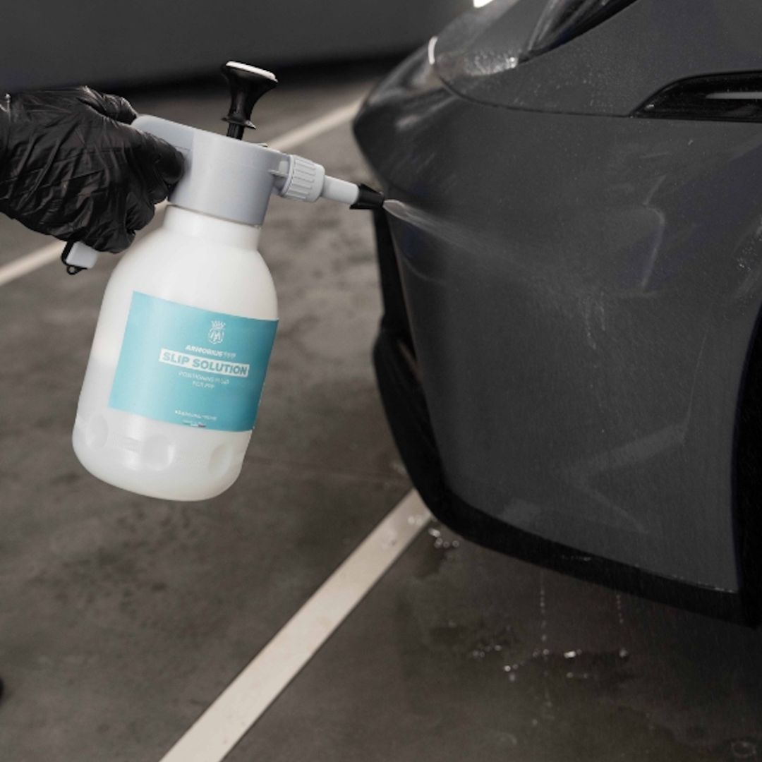 A gloved hand holds a white spray bottle labeled 'PREMIER PPF SLIP SOLUTION' and sprays a dark grey car body in a garage setting.