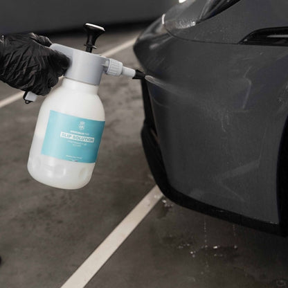 A gloved hand holds a white spray bottle labeled 'PREMIER PPF SLIP SOLUTION' and sprays a dark grey car body in a garage setting.