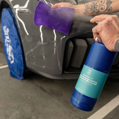 A person sprays a blue and teal bottle labeled 'PPF Surface Reset' onto a purple microfibre cloth, which is then wiped onto a dark gray car body. A blue protective cover is draped over the car wheel.