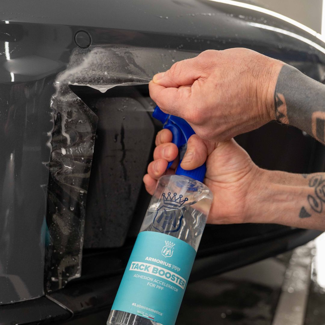 A close-up of a person's tattooed hands applying Labocosmetica ARMORIUS PPF Tack Booster to the front bumper of a black car