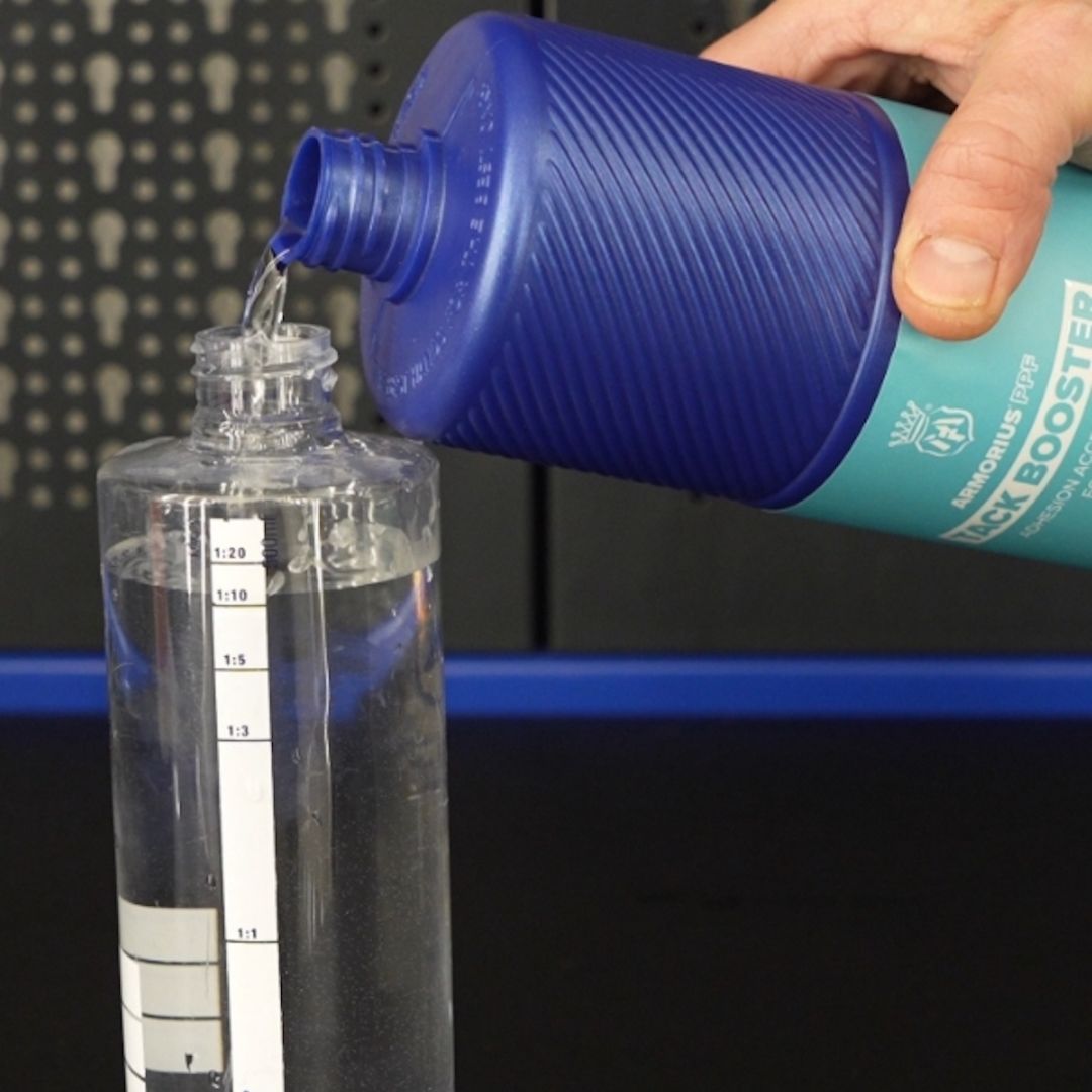 A hand pouring liquid from a blue and teal bottle labeled 'Tack BOOSTER' into a clear measuring container with printed measurement markings.