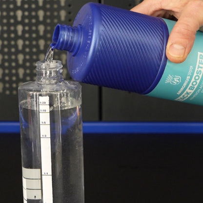 A hand pouring liquid from a blue and teal bottle labeled 'Tack BOOSTER' into a clear measuring container with printed measurement markings.