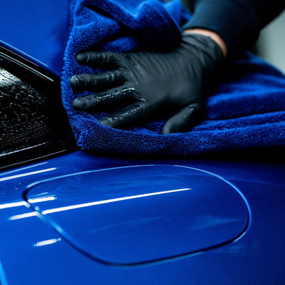 A close-up of a hand wearing a black glove, wiping a glossy blue car body with a textured blue Labocosmetica drying towel.
