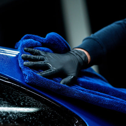 A close-up of a hand wearing a black glove, wiping a glossy blue car body with a textured blue Labocosmetica drying towel.
