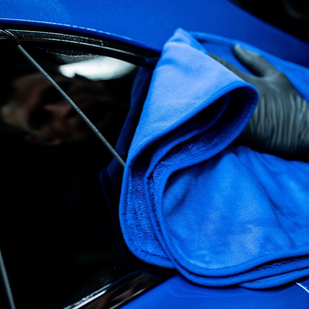 A close-up of a hand wearing a black glove, wiping a glossy blue car body with a textured blue Labocosmetica drying towel.
