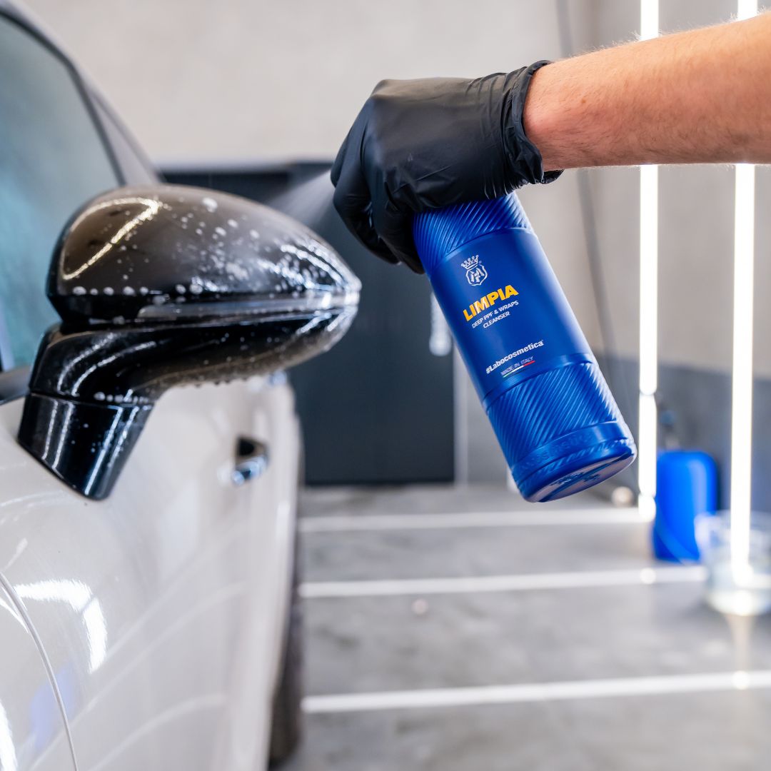 Person cleaning a PPF car mirror with a blue bottle labeled 'Limpia' in a garage setting.