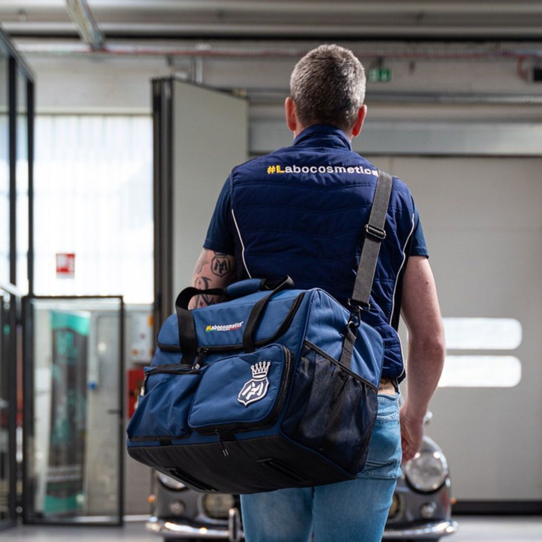 Man wearing a blue shirt with 'Labocoetmetica' branding, carrying a matching blue duffel bag in an indoor setting.