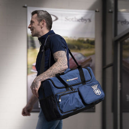 Man carrying a large blue duffel bag with a logo in an indoor setting. Labocosmetica Multi Tool Bag – Large Detailing & Polisher Bag (Size L)