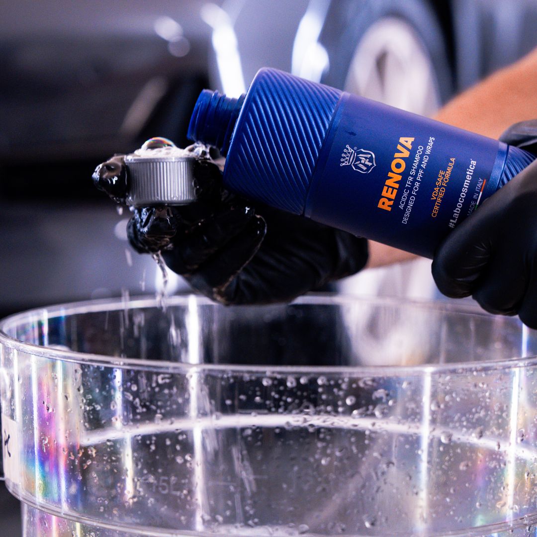 Hand holding a blue bottle labeled Laboosmetica Renova over a clear bucket with a blurred car in the background