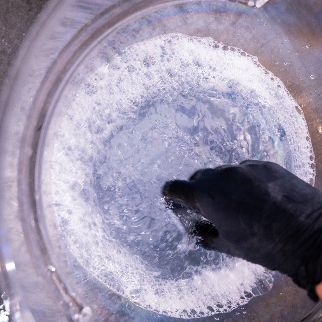 Hand wearing a black glove stirring a pot of bubbling liquid with foam.