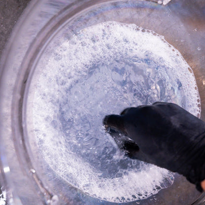 Hand wearing a black glove stirring a pot of bubbling liquid with foam.