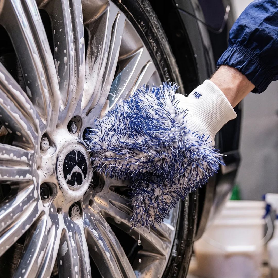 A close-up of a hand washing a silver car wheel with soapy water, wearing a blue and white microfibre cleaning glove by Labocosmetica