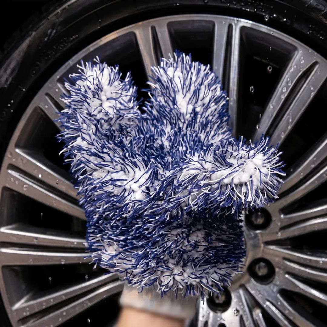 A close-up of a hand washing a silver car wheel with soapy water, wearing a blue and white microfibre cleaning glove by Labocosmetica