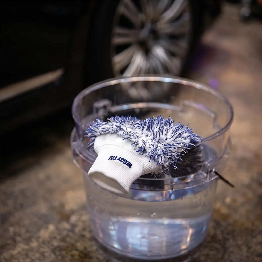 A car wash mitt glove from Labocosmetica with Reggy Fox, featuring a navy blue and white shaggy scrubber top on a white knit cuff hanging over the edge of a see through wash bucket