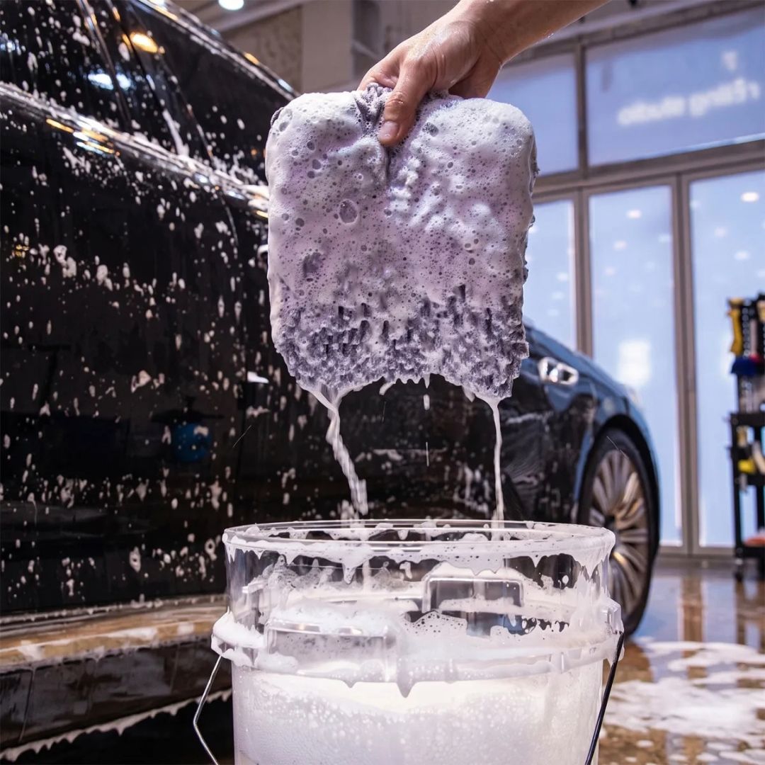 A hand holds a thick, soapy, foam-covered sponge above a bucket filled with white cleaning soap, with a wet black car and a car wash shop visible in the background.