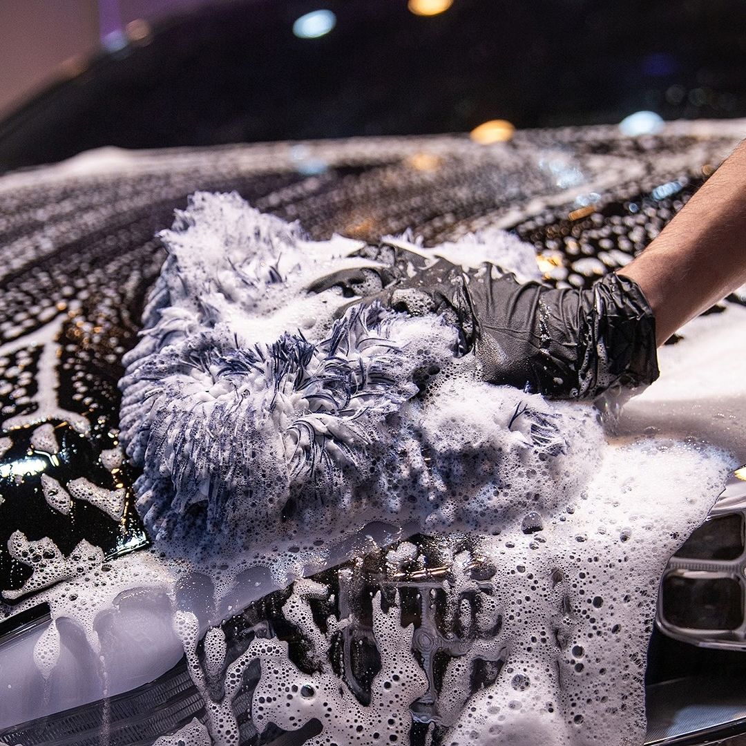 A close-up action shot of a person wearing a black glove and using a thick, high-pile blue and white microfiber wash mitt to scrub a black car hood covered in soapy foam.