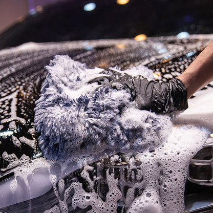 A close-up action shot of a person wearing a black glove and using a thick, high-pile blue and white microfiber wash mitt to scrub a black car hood covered in soapy foam.