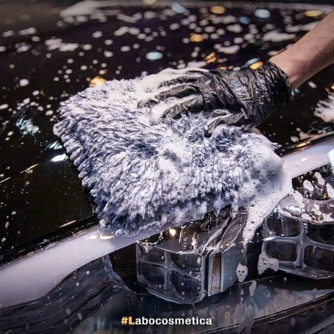 A close-up action shot of a hand wearing a black textured glove holding a fluffy gray microfiber wash cloth covered in soapy foam, scrubbing a glossy black car surface. The hashtag #Labocosmetica is visible at the bottom.