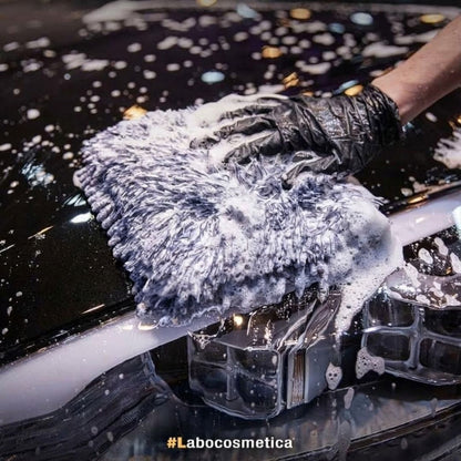 A close-up action shot of a hand wearing a black textured glove holding a fluffy gray microfiber wash cloth covered in soapy foam, scrubbing a glossy black car surface. The hashtag #Labocosmetica is visible at the bottom.