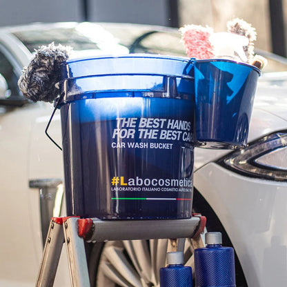 A blue car wash bucket with a fuzzy brush on the handle, set on a small step stool next to a white car. The bucket has white text reading 'THE BEST HANDS FOR THE BEST CAR WASH BUCKET' and '#Labocosmetica'.