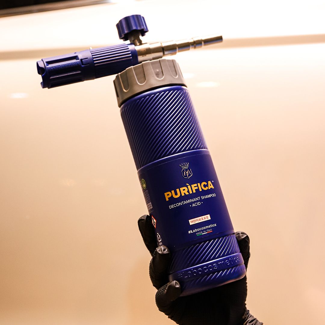 A person wearing a black glove holds a Purifica Contaminant Removal + Acids 400ml portable Labocosmetica bottle against a warm beige background.