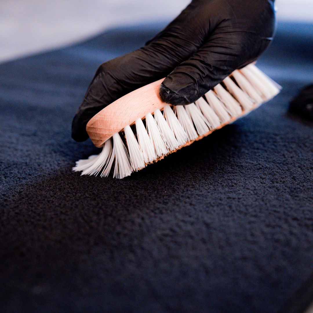 Hand wearing a black glove holding a brush against a car mat