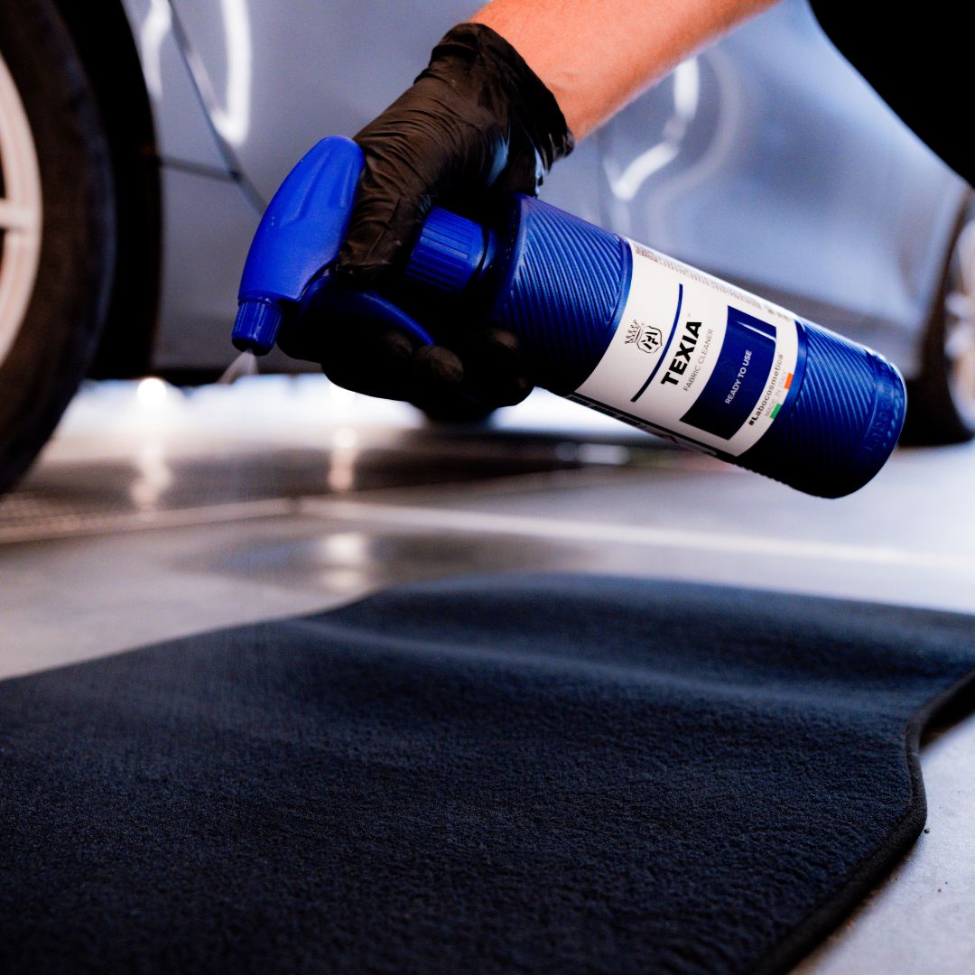 Person holding a blue spray bottle labeled 'Texia' next to a car floor mat.