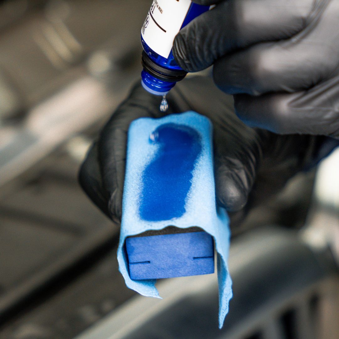 Person applying ceramic coating liquid from a bottle onto a blue sponge with a blurred vehicle interior background.