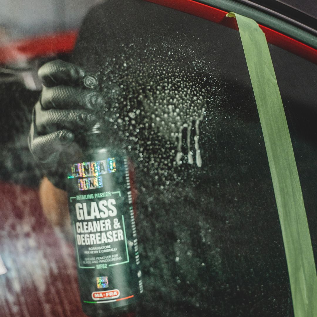 Hand holding a bottle of glass cleaner and degreaser on a car window with water droplets.