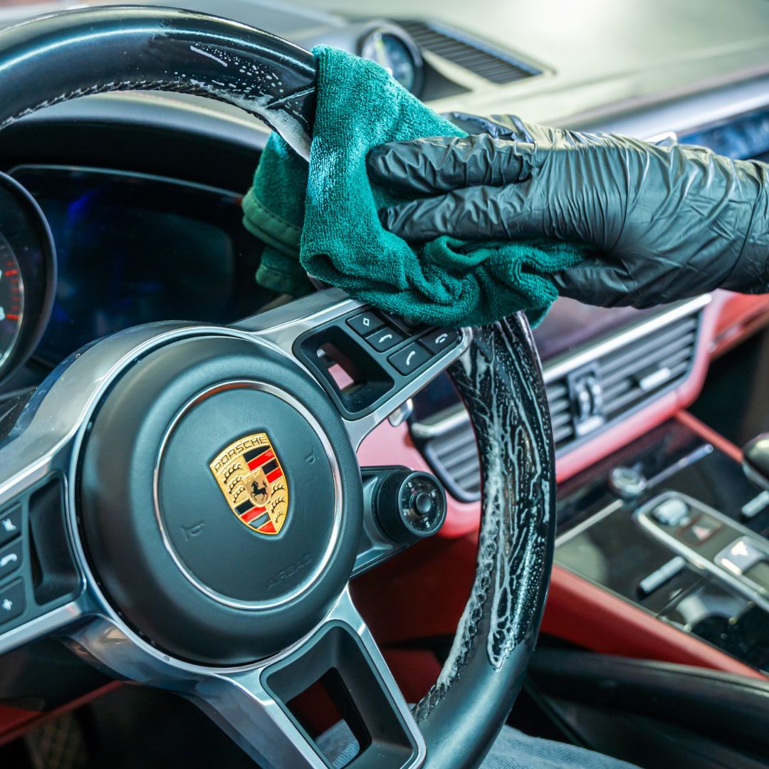 Person cleaning a Porsche steering wheel with a green cloth inside a car.