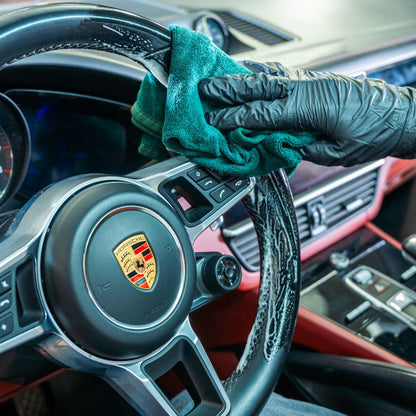Person cleaning a Porsche steering wheel with a green cloth inside a car.