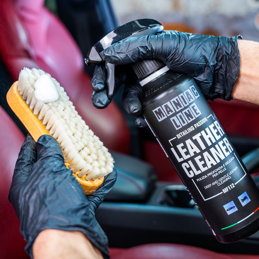Person holding Maniac leather cleaner bottle and brush with black gloves in a car interior.