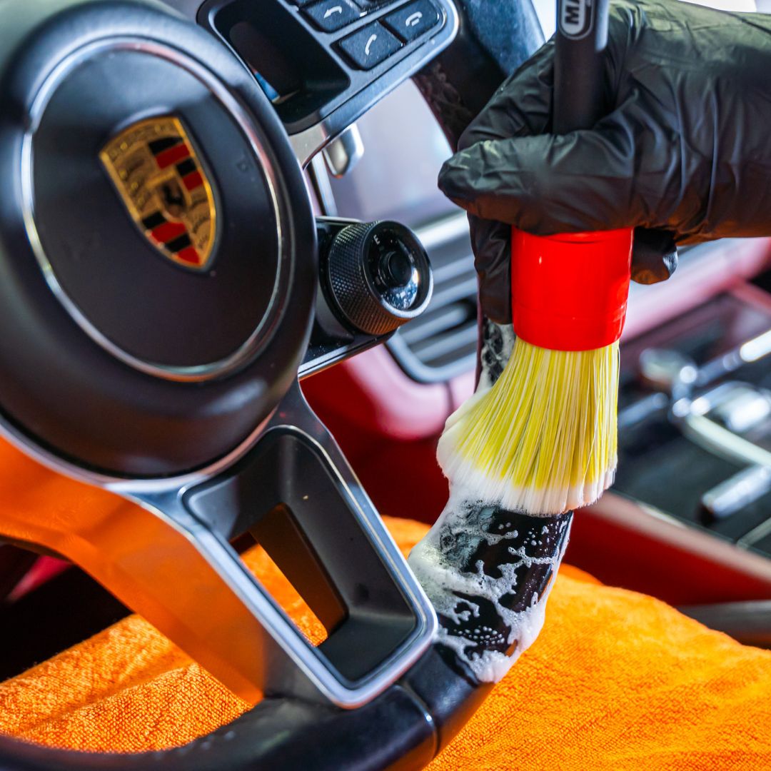 Person cleaning a Porsche steering wheel with a brush and orange towel.