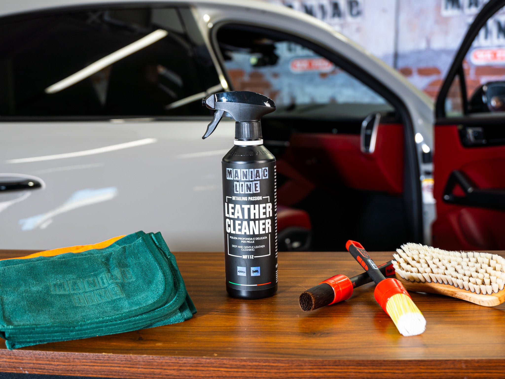 Maniac Line leather cleaner spray bottle, towel, brush, and tools on a wooden surface with a Porsche Panamera in the background.