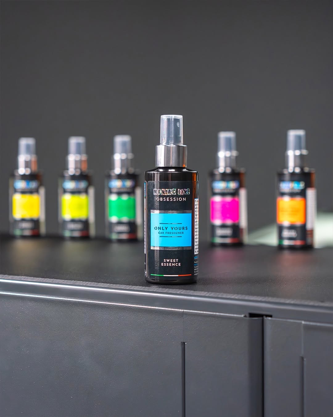 A bottle of Maniac Line Obsession air freshener spray stands in sharp focus on a dark grey surface, with five other colourful spray bottles blurred in the background against a dark grey wall.