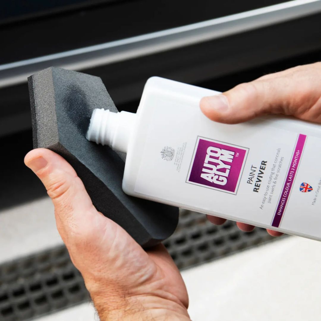 Person holding a bottle of Autoglym Paint Reviver with a precision foam applicator against a car door.
