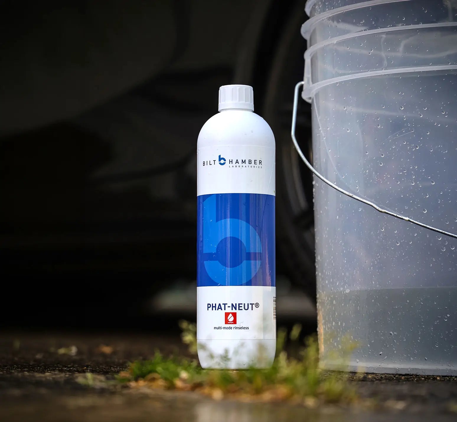 A bottle of Bilt Hamber Phat-Neut cleaner stands on wet pavement next to a translucent plastic bucket, with blurred car wheels and grass in the background.