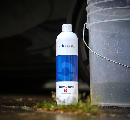 A bottle of Bilt Hamber Phat-Neut cleaner stands on wet pavement next to a translucent plastic bucket, with blurred car wheels and grass in the background.