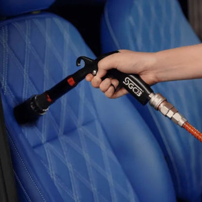 A hand holding a black SCGB air nozzle connected to an orange hose, cleaning a blue quilted car seat.