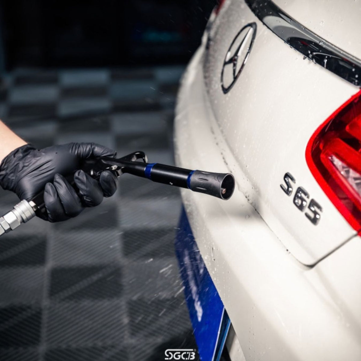 A close-up action shot of a person wearing a black glove and using an SGCB air gun to dry a  white Mercedes-Benz S65 car. The background shows a dark checkered floor, and the Mercedes-Benz logo and 'S65' model badge are visible on the car.
