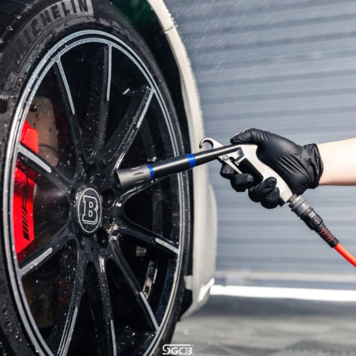A close-up of a black Brabus Mercedes wheel being dried with an SGCB Air gun held by a person wearing a black glove. The wheel has a silver and black multi-spoke design with a visible red brake caliper and a circular 'B' logo in the center.