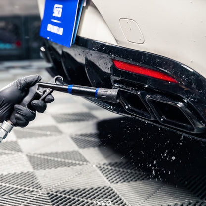 A close-up action shot of a person wearing a black glove using an SGCB Tornador air gun to dry the rear diffuser and exhaust area of a white mercedes. The car has a black rear diffuser with dual exhaust outlets and a red taillight, set on a gray checkered floor.