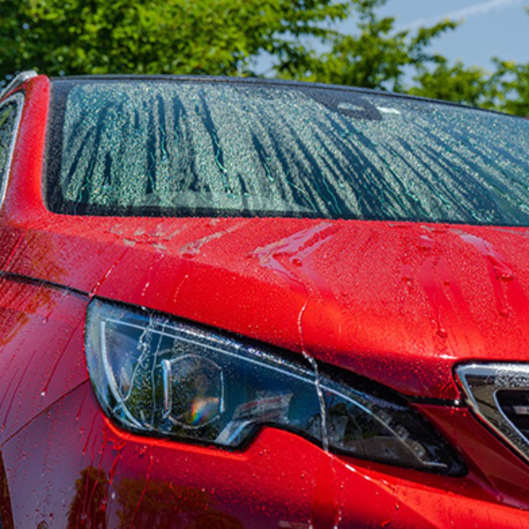 Close-up of a red car's front with water droplets on the windshield and headlight.