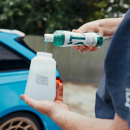 Person pouring a liquid from a bottle labeled 'stjarnagloss Bubblor into another container with a blue car in the background.