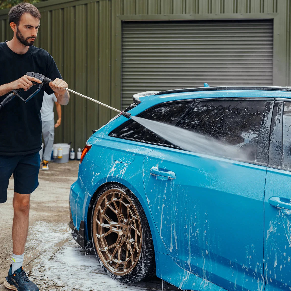 Man washing a blue car with a pressure washer in front of a green building.