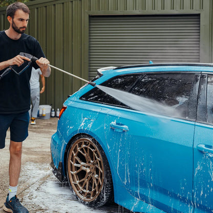 Man washing a blue car with a pressure washer in front of a green building.