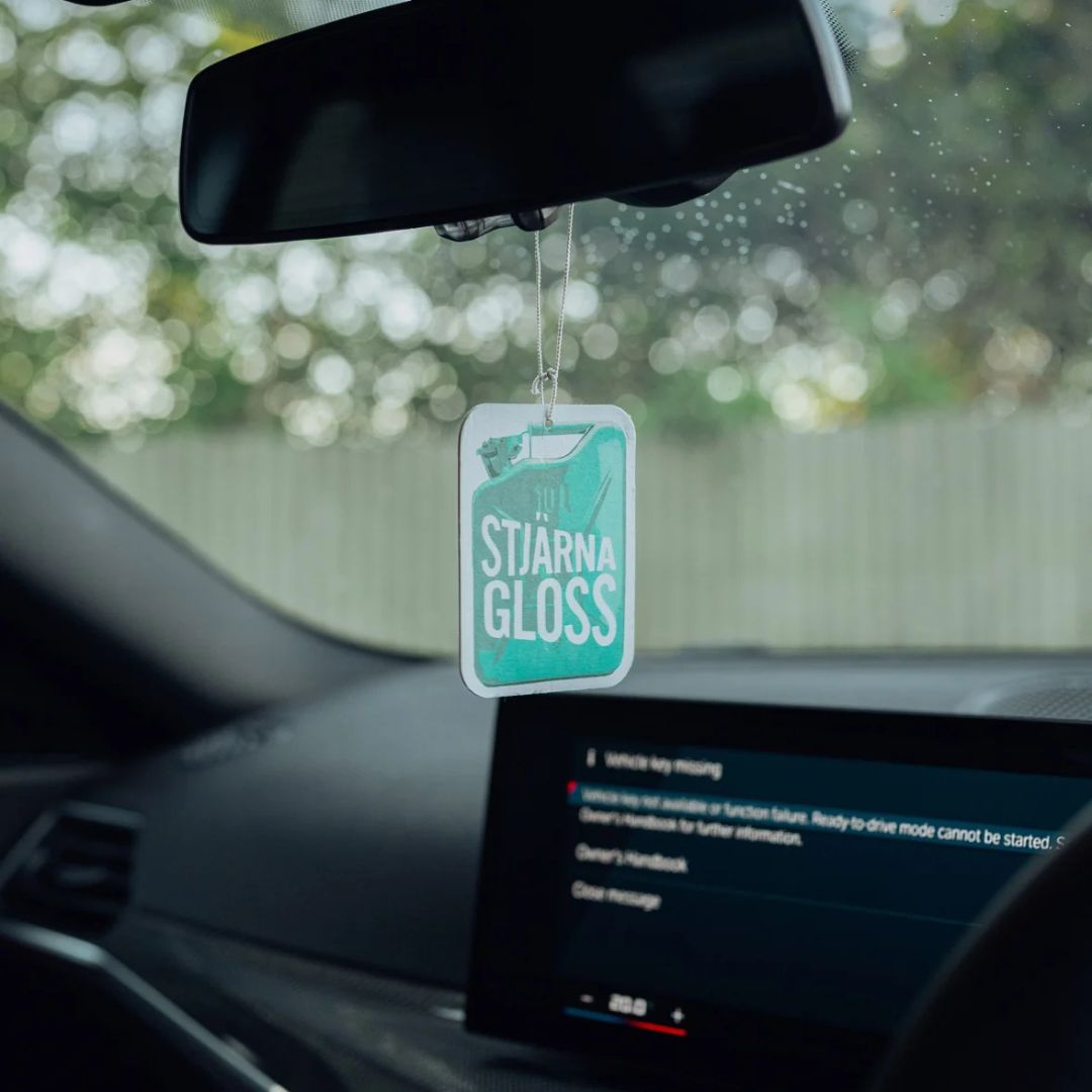 A Stjärnagloss car air freshener hanging from a car's rearview mirror. The rectangular, cloud-shaped air freshener is light teal with white text reading 'STJÄRNA GLOSS' and a small illustration of a car.
