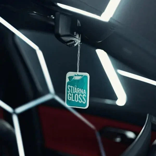 A car interior air freshener tag labeled 'STJÄRNAGLOSS' hanging from the rearview mirror, set against a dark car cabin with geometric LED lighting.