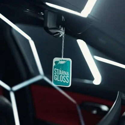 A car interior air freshener tag labeled 'STJÄRNAGLOSS' hanging from the rearview mirror, set against a dark car cabin with geometric LED lighting.